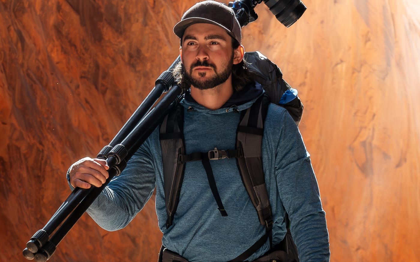 Forest Barkdoll-Weil carrying a camera and tripod on his shoulder, standing in a desert canyon with orange rock formations, wearing outdoor gear.