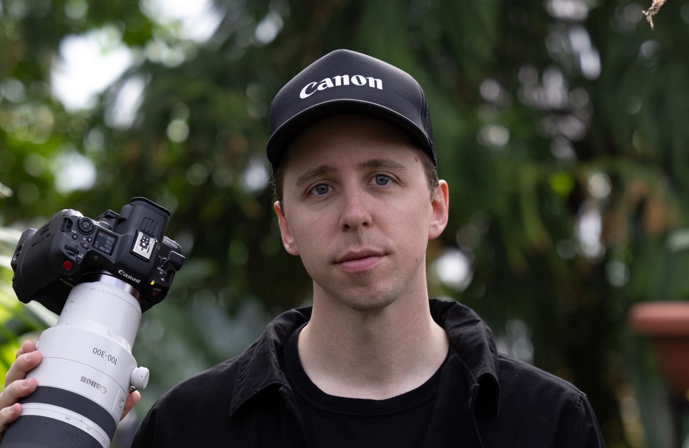 Portrait of Ben Haggarty holding a Canon camera with a large telephoto lens, wearing a black Canon cap and jacket, set against a blurred outdoor background.