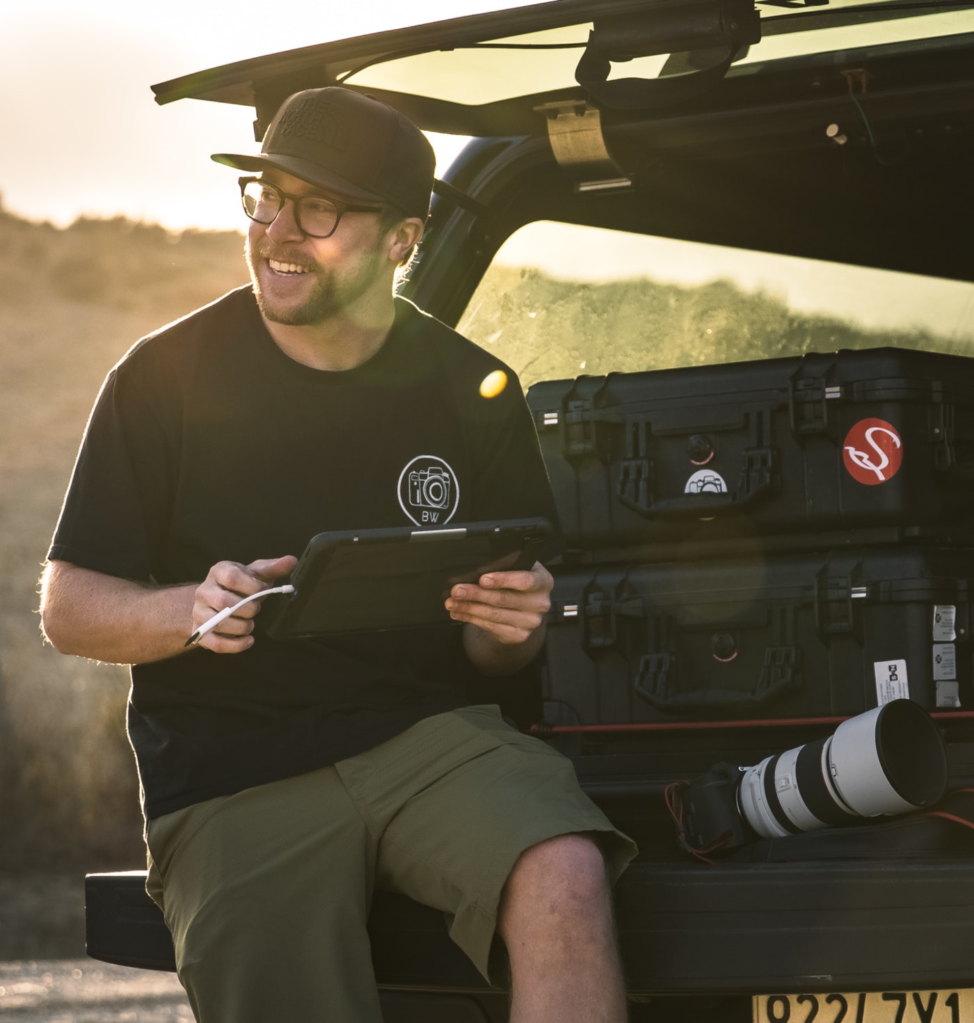 A smiling Brian Walker sits at the back of a vehicle, holding a tablet and surrounded by photography gear, including a camera with a large telephoto lens, during a sunny outdoor shoot.