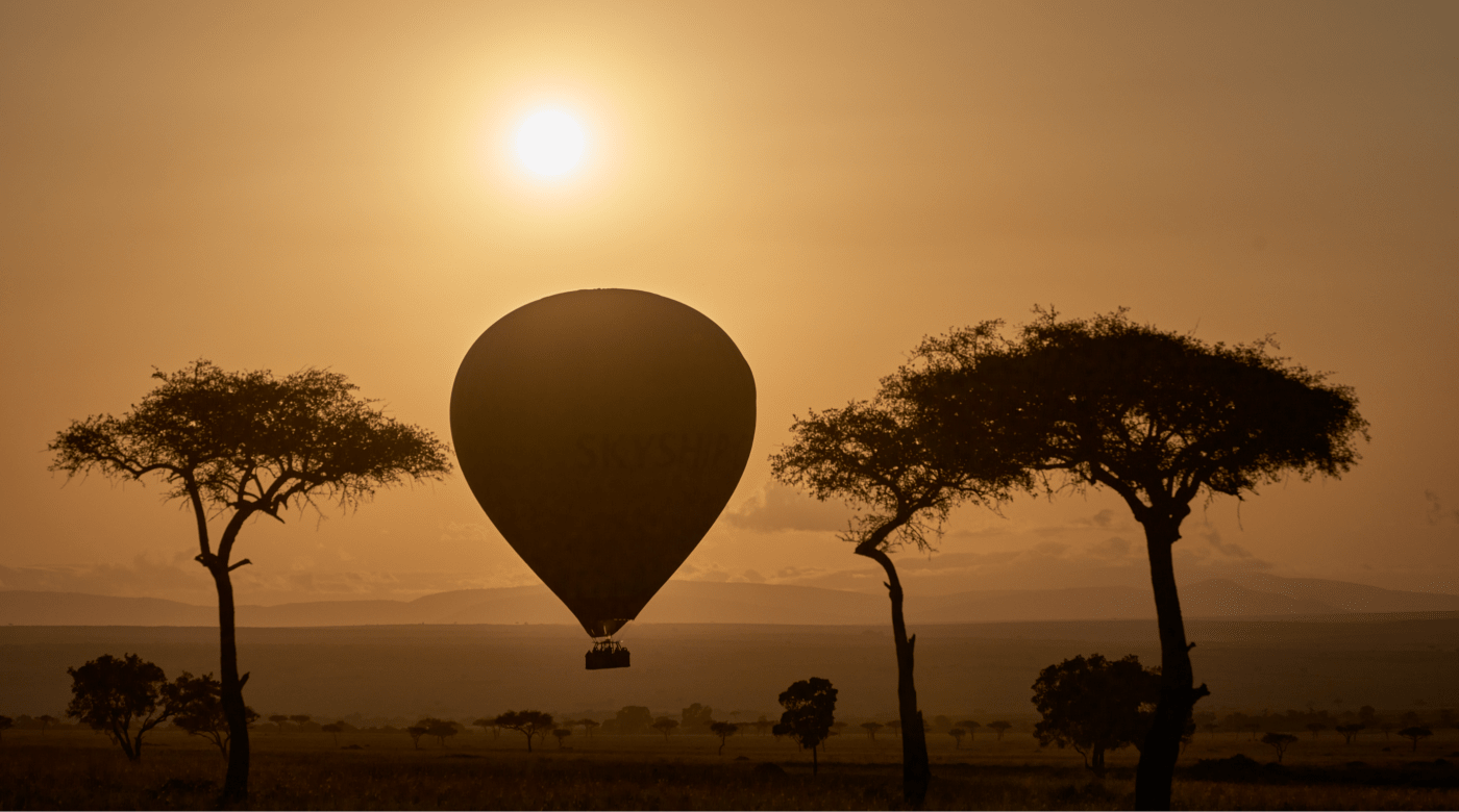 Silhouette of a hot air balloon with the text 'SKYSHIP' floating at sunset between two trees in a savanna landscape, photographed by McNally.