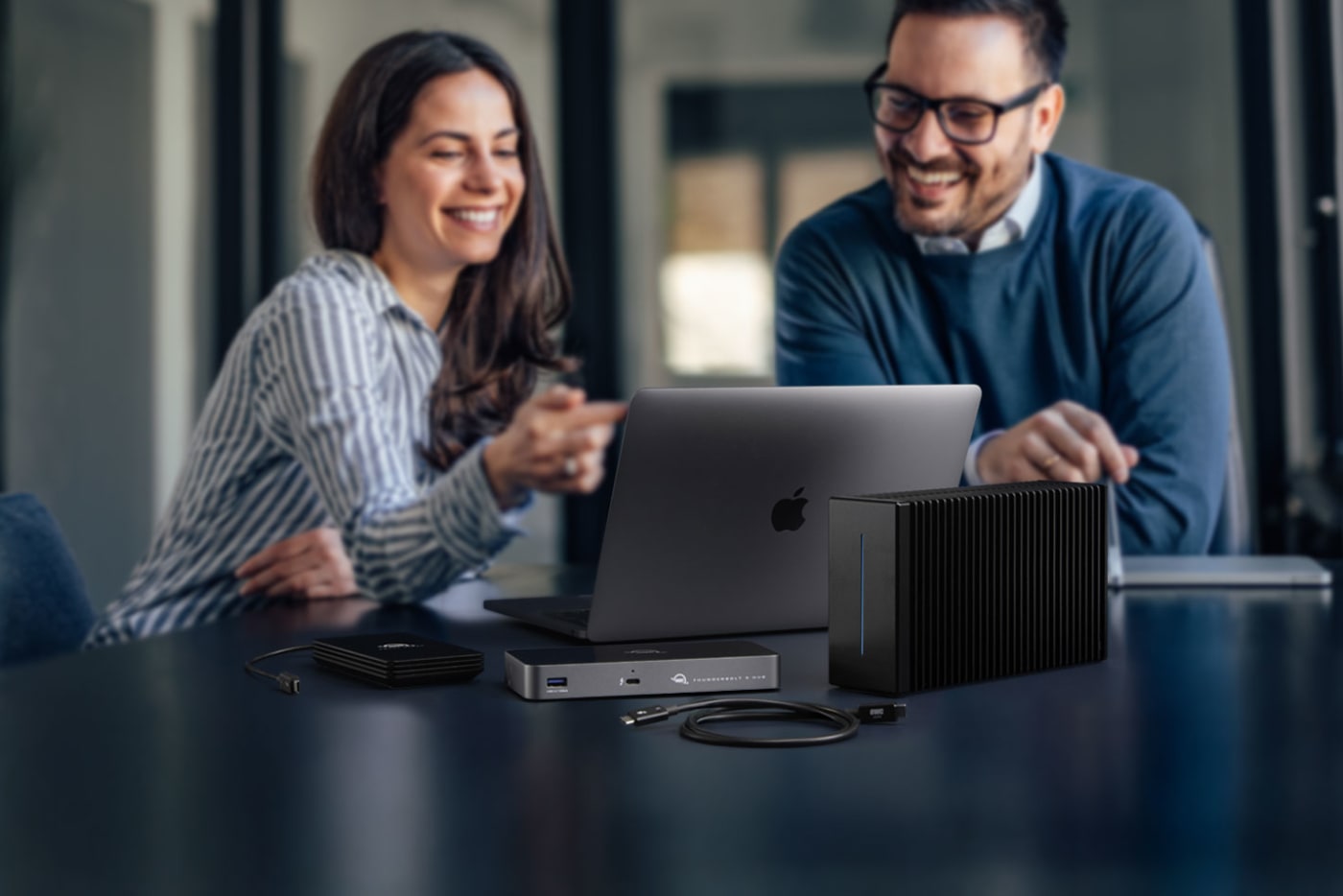 Two people looking at a MacBook Pro on desktop with OWC Thunderbolt 5 Hub and OWC Envoy Ultra