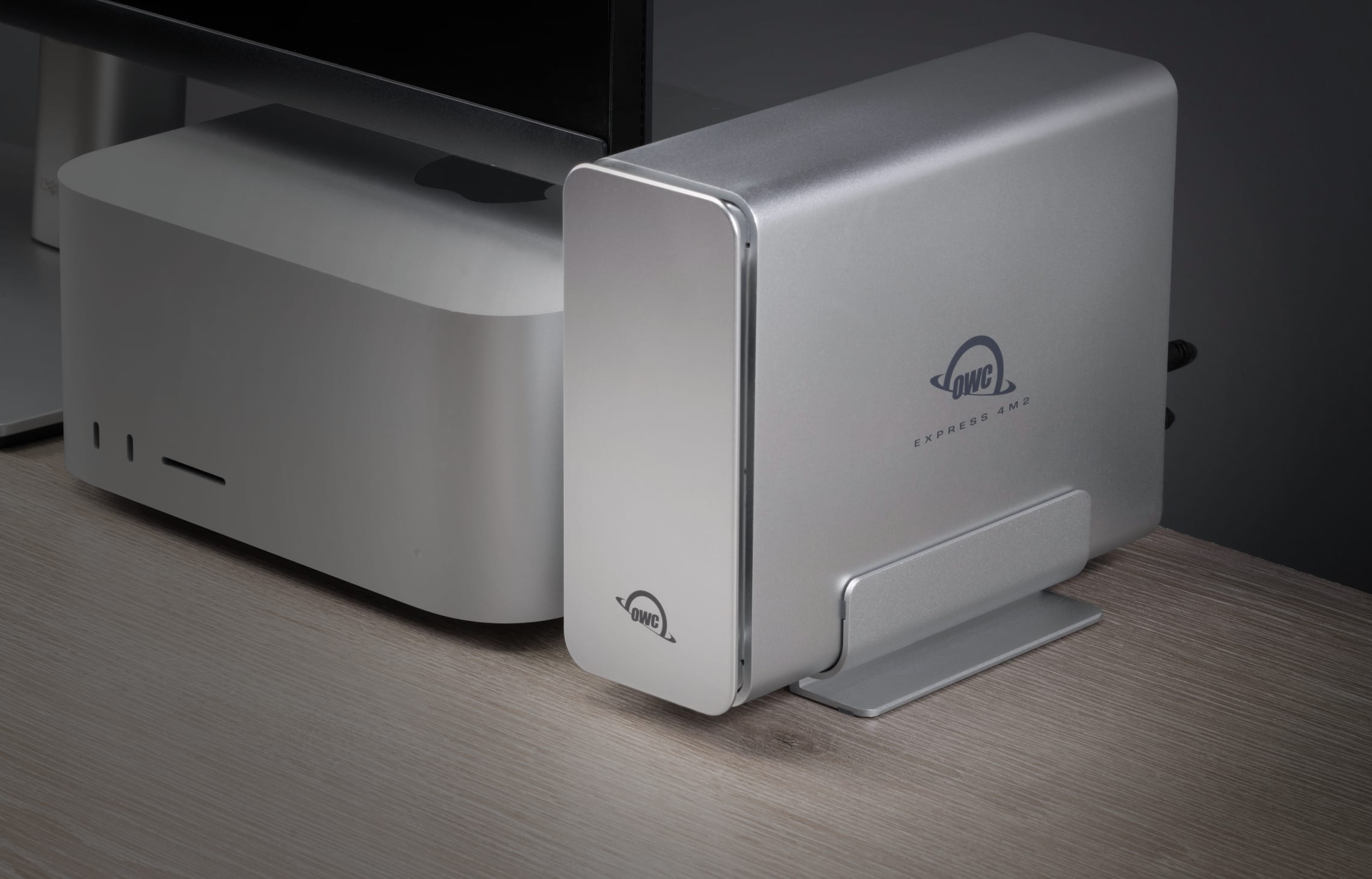 OWC Express 4M2 USB4 NVMe SSD Enclosure placed vertically next to a Mac Studio and monitor, showcasing its compact, space-saving design and professional desktop integration.