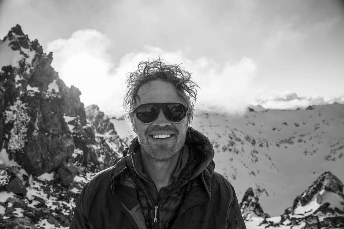 A black-and-white portrait of Adam Clark smiling in front of a mountainous, snow-covered landscape. He is wearing sunglasses and a warm jacket, with the bright sun peeking through the clouds in the background, casting a soft glow over the rugged terrain.