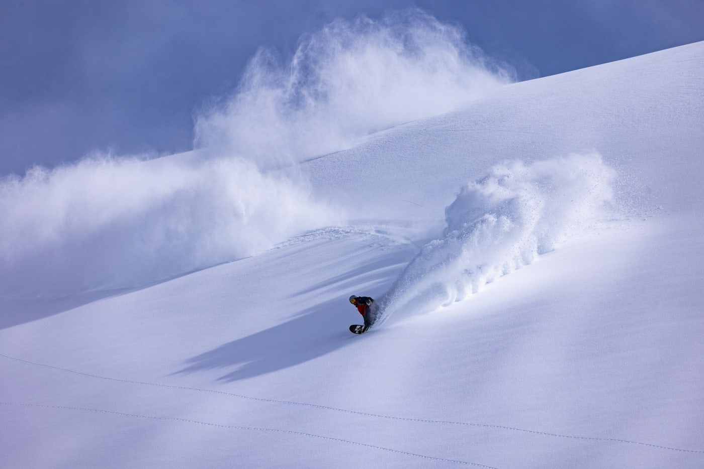 A lone snowboarder carves down a pristine snow-covered mountain, leaving a trail of powder in their wake under a clear blue sky.