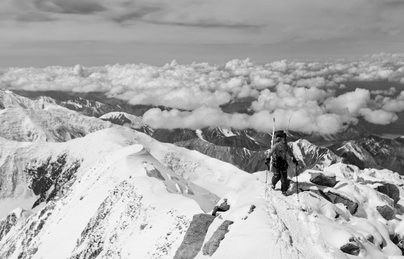 A climber stands on the edge of a snow-covered mountain ridge, overlooking a sea of clouds, with rugged peaks extending into the horizon.