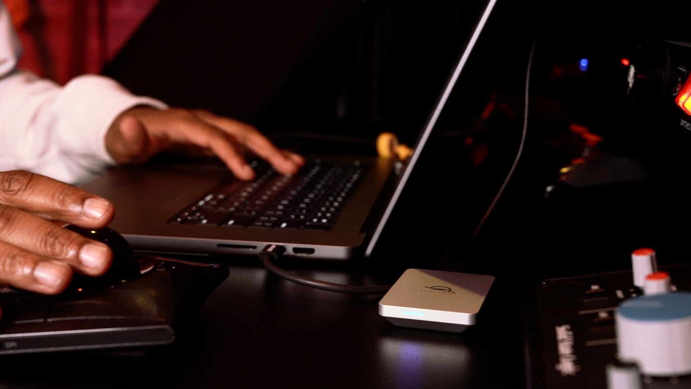 Adrian Porter's hands working on a trackball mouse and laptop, with an OWC Elektron and audio equipment visible on the desk.