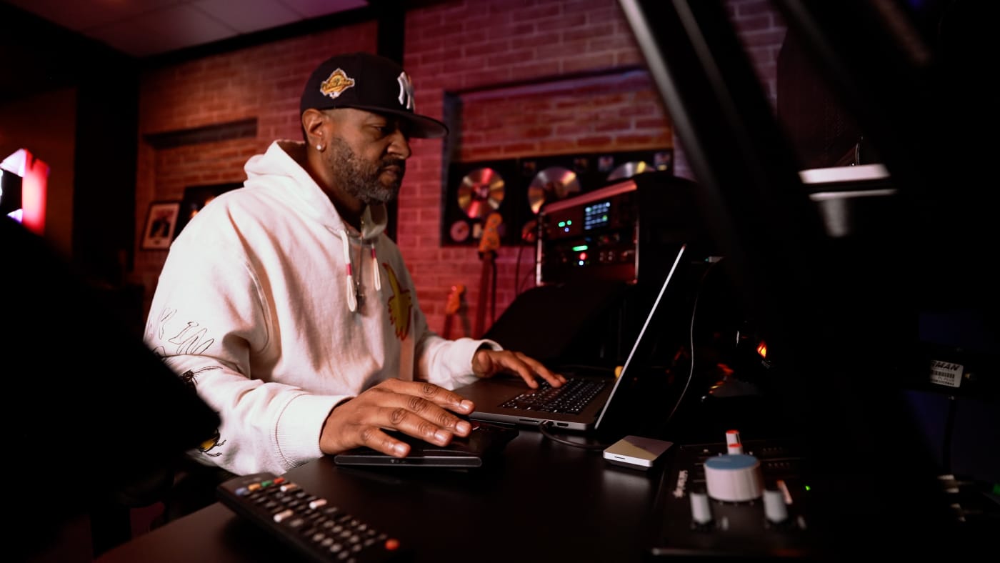 Adrian Porter working at a desk with audio equipment, using a laptop in a studio with gold records on the wall in the background.