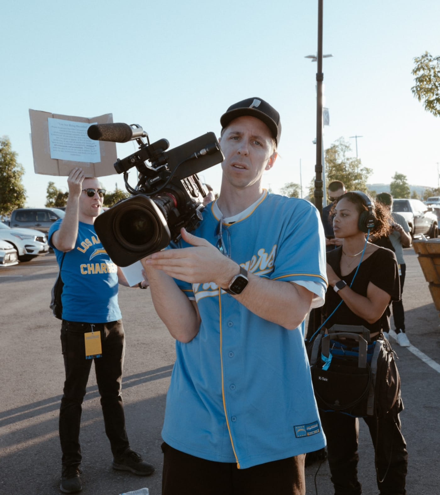 Ben Haggarty holding a professional video camera while wearing a Los Angeles Chargers jersey and a black cap, with a film crew working behind him in an outdoor setting.