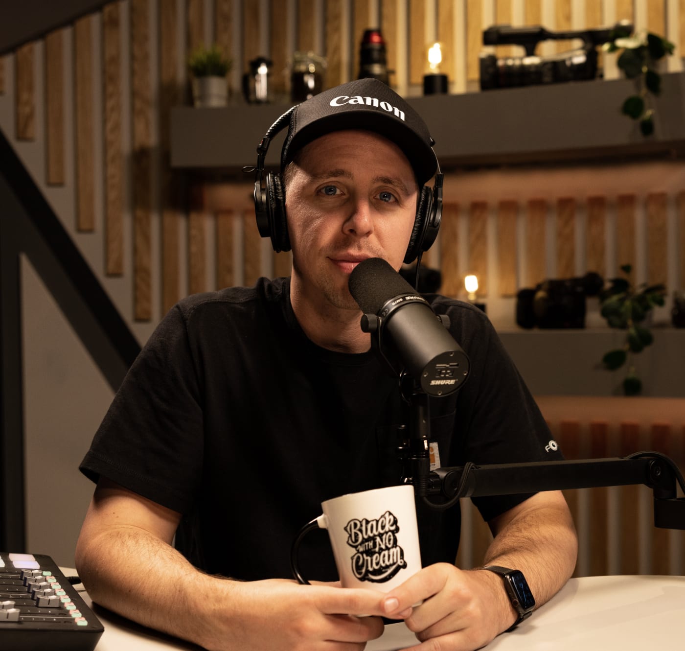 Ben Haggarty hosting a podcast, speaking into a microphone while wearing headphones and holding a 'Black With No Cream' mug, in a studio with camera equipment in the background.