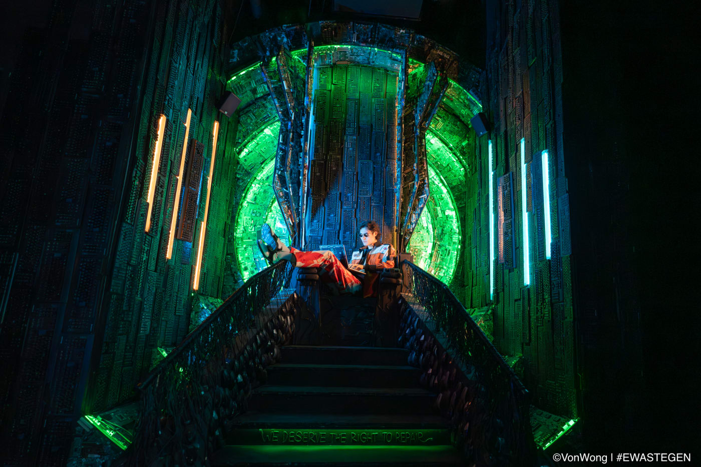 A person sitting confidently on a futuristic throne made of circuit boards, lit in vibrant green and blue hues, evoking a sci-fi atmosphere.