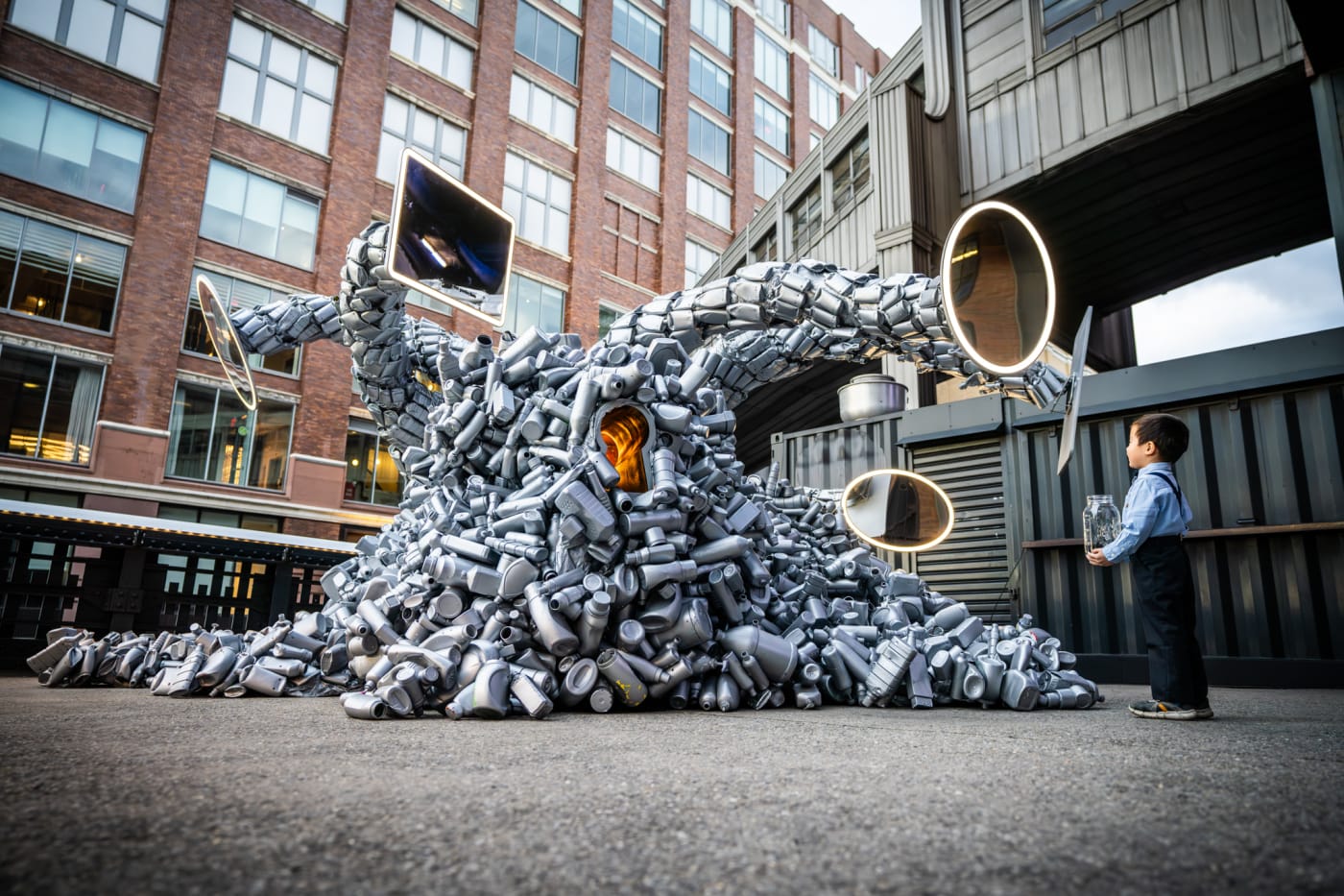 A young child standing in awe before a massive sculpture made from repurposed metal objects, featuring tentacle-like appendages with mirrors at their ends. The installation stands against an urban backdrop, creating a sense of wonder.