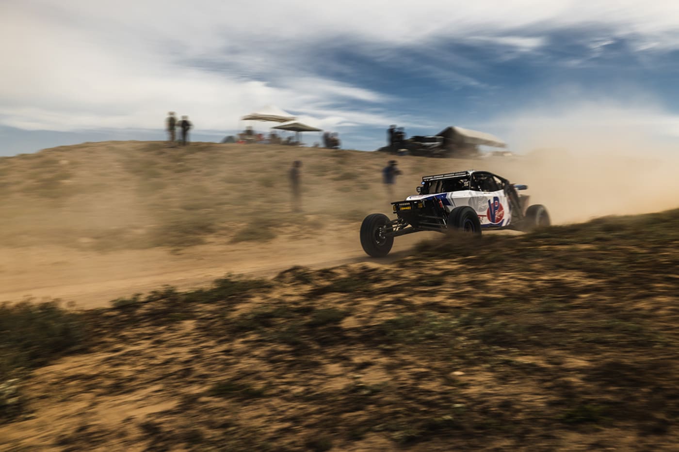 A Baja off-road racing vehicle kicking up dust while speeding across a desert landscape, with spectators watching from a hill in the distance.