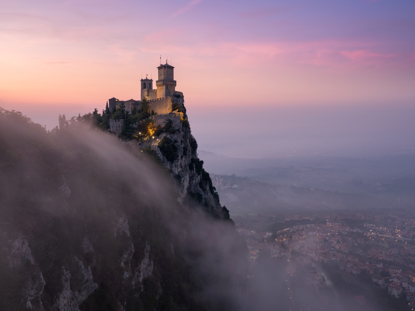 Photograph by Elia Locardi of a medieval castle perched on a cliff in San Marino at sunrise, with mist rolling over the landscape and a pink and purple sky.
