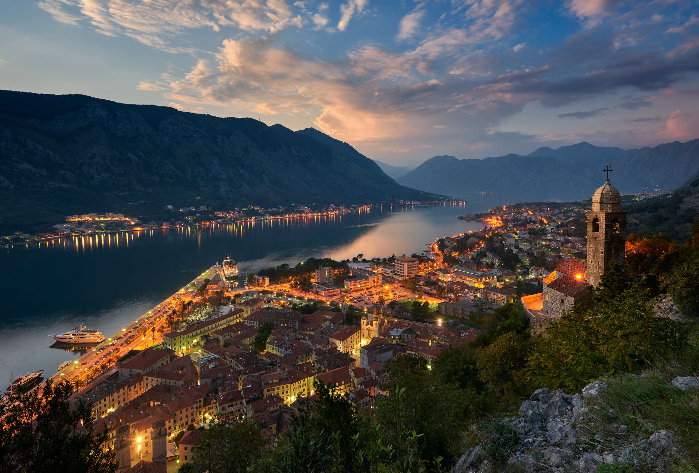 Photograph by Elia Locardi of Kotor Bay in Montenegro at dusk, with illuminated streets and buildings along the coastline, surrounded by mountains and a view of a church tower in the foreground.