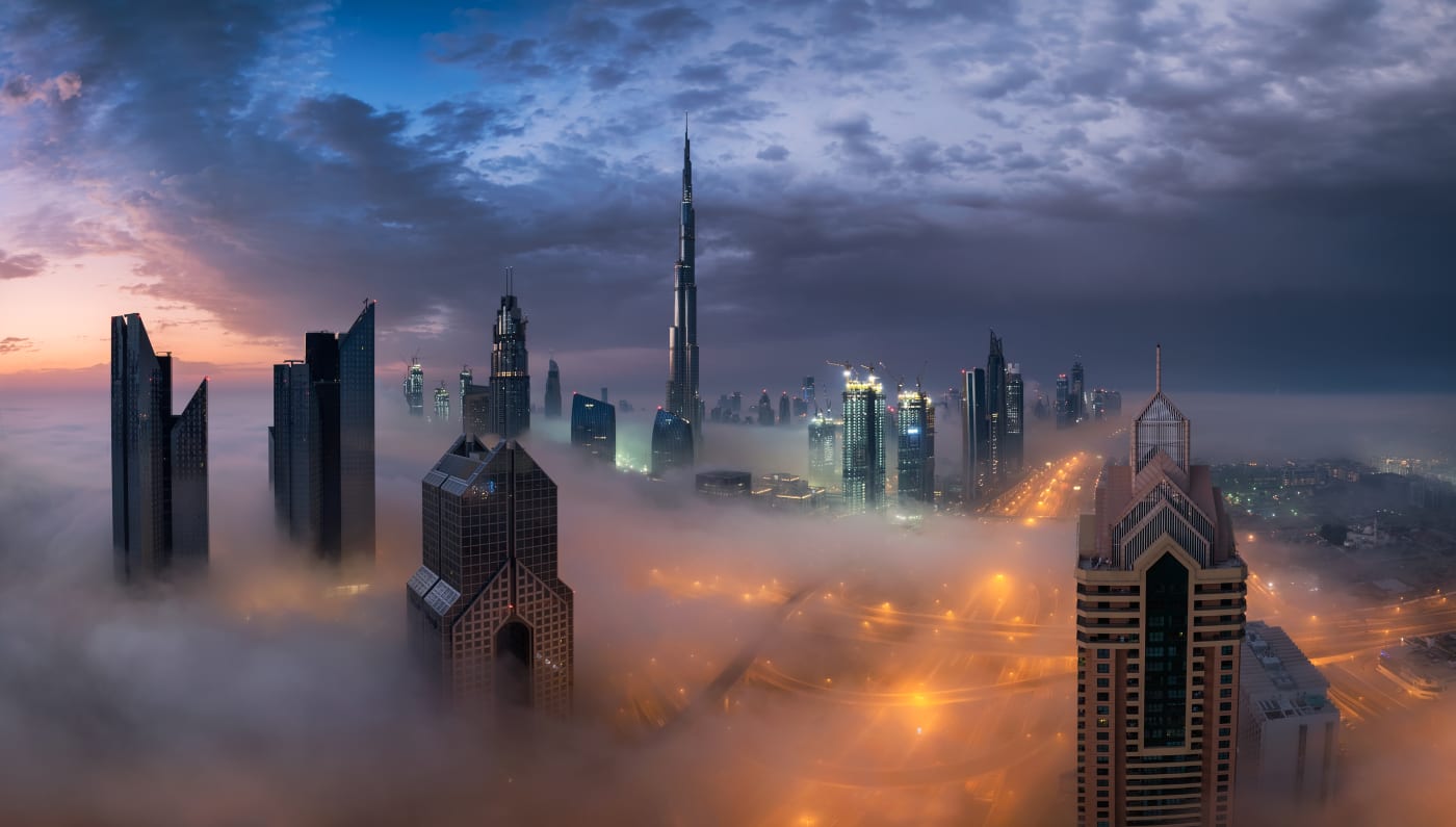 Photograph by Elia Locardi of the Dubai skyline at sunrise, with tall skyscrapers, including the Burj Khalifa, emerging through a dense layer of fog and illuminated streets below.