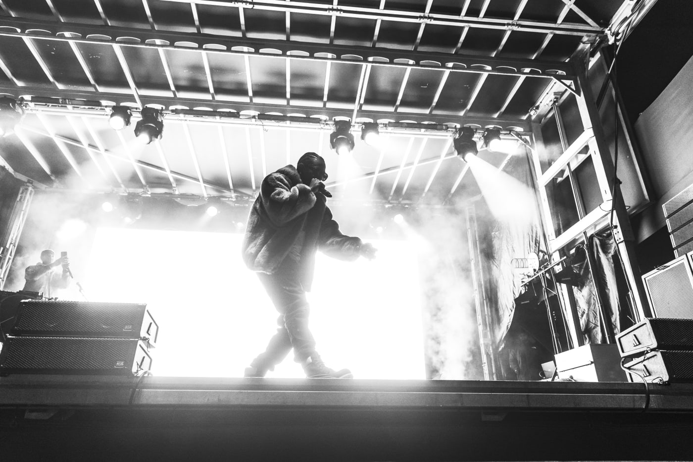 A black and white image of a performer on stage under bright lights, mid-performance, with fog effects around.