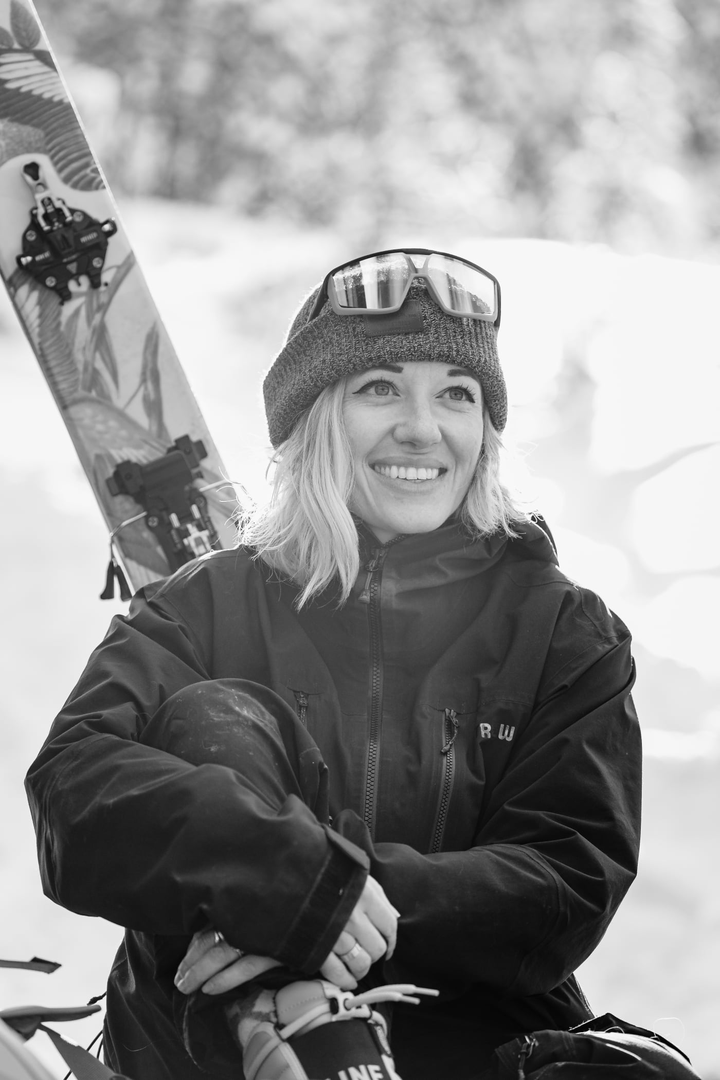 Black and white portrait of Emily Tidwell smiling while sitting in snow gear, with ski goggles on her beanie and skis in the background.