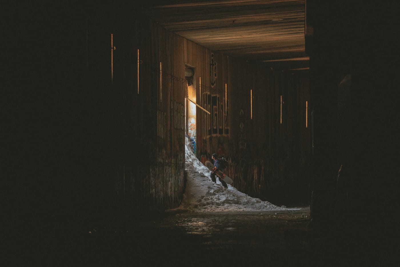 A snowboarder climbing a snowy slope inside a tunnel with graffiti-covered walls, light coming from the exit.