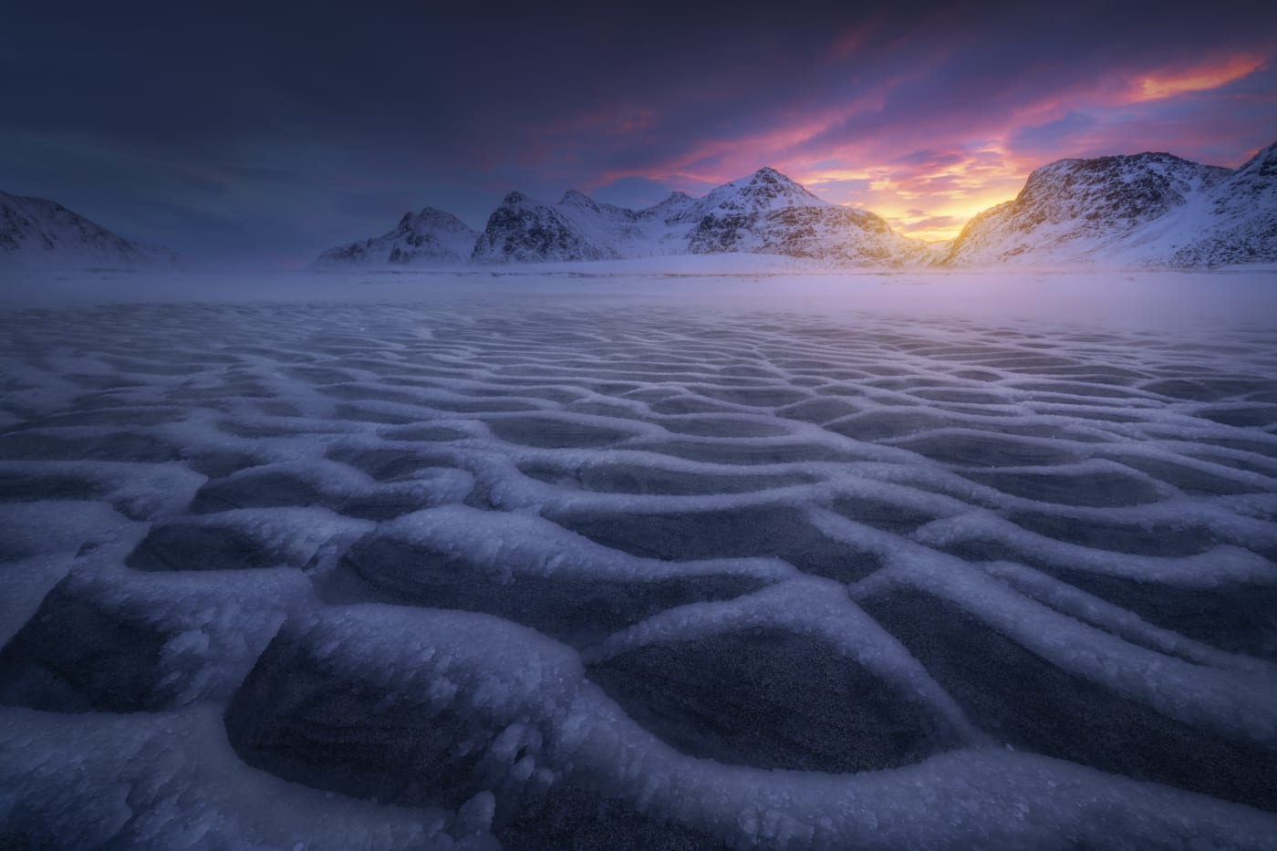 Photograph by Forest Barkdoll-Weil of a snow-covered field with textured ice patterns, leading to distant snow-capped mountains at sunrise with colorful clouds in the sky.