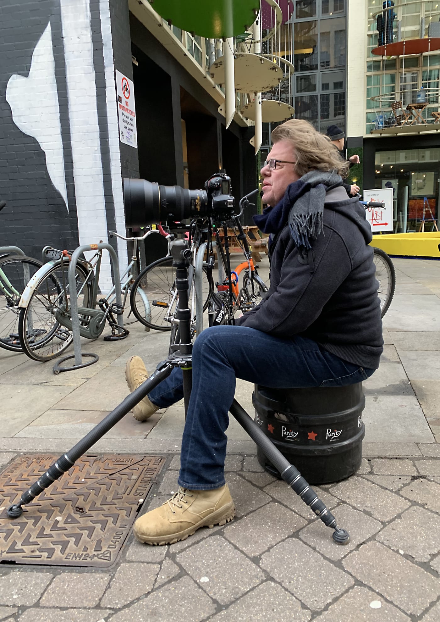 Photographer Joe McNally seated on a barrel on a city street, operating a large camera with a telephoto lens mounted on a tripod, surrounded by bicycles and urban scenery.