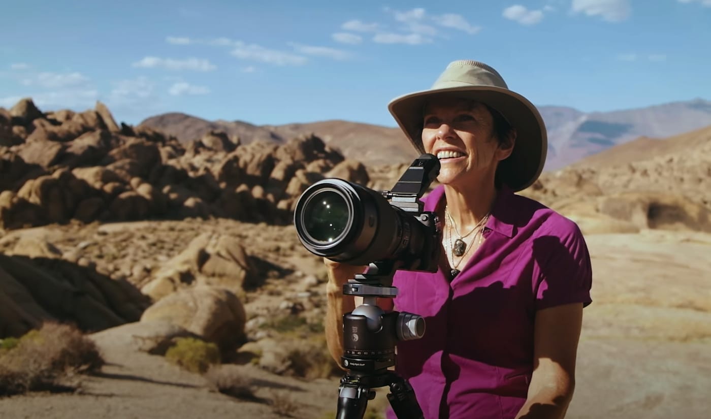 Karen Hutton smiling while holding a camera on a tripod in a desert landscape, wearing a wide-brimmed hat and purple shirt under clear blue skies.