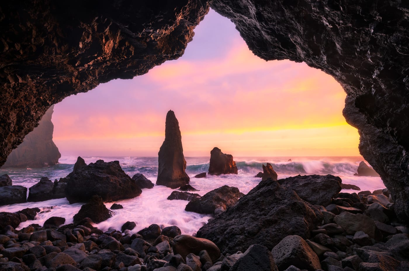 A vibrant sunset view from within a sea cave, with towering sea stacks in the crashing waves, casting warm light across the rocky shoreline.
