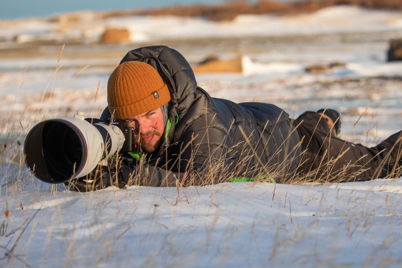 A photographer, Nate Luebbe, lying on the snowy ground in a puffer jacket and beanie, aiming through a telephoto camera lens during an outdoor shoot.