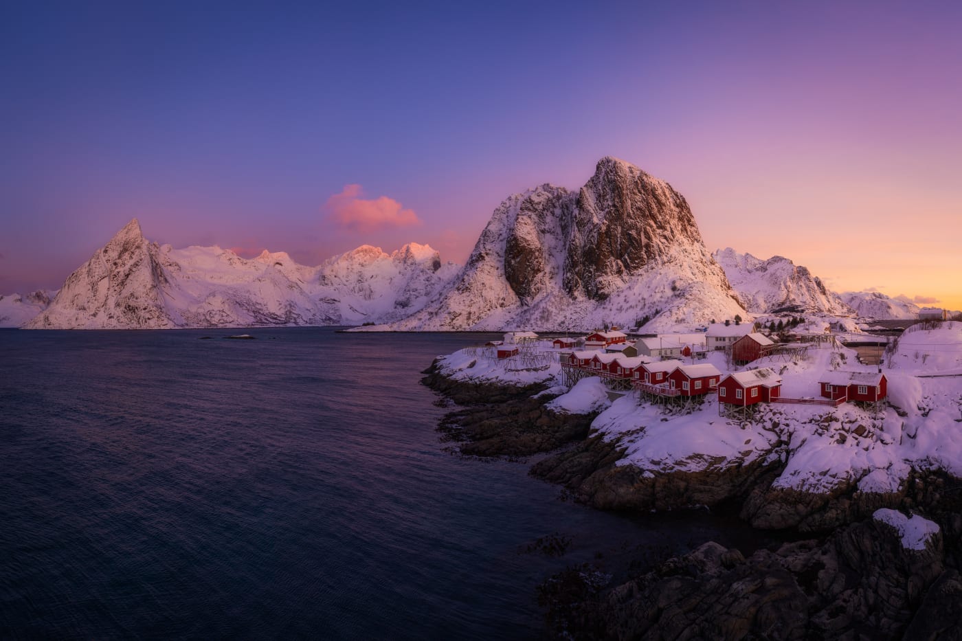 Image of a coastal town nestled by snow-capped mountains during a purple sunset, with small red cabins along the shoreline.