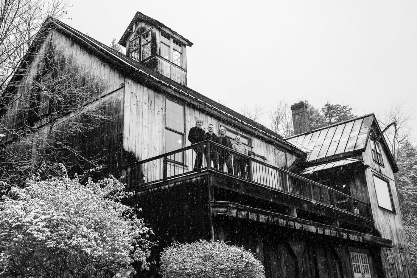 A black and white image of a band standing on a wooden house balcony, with snow gently falling and barren trees surrounding the scene.
