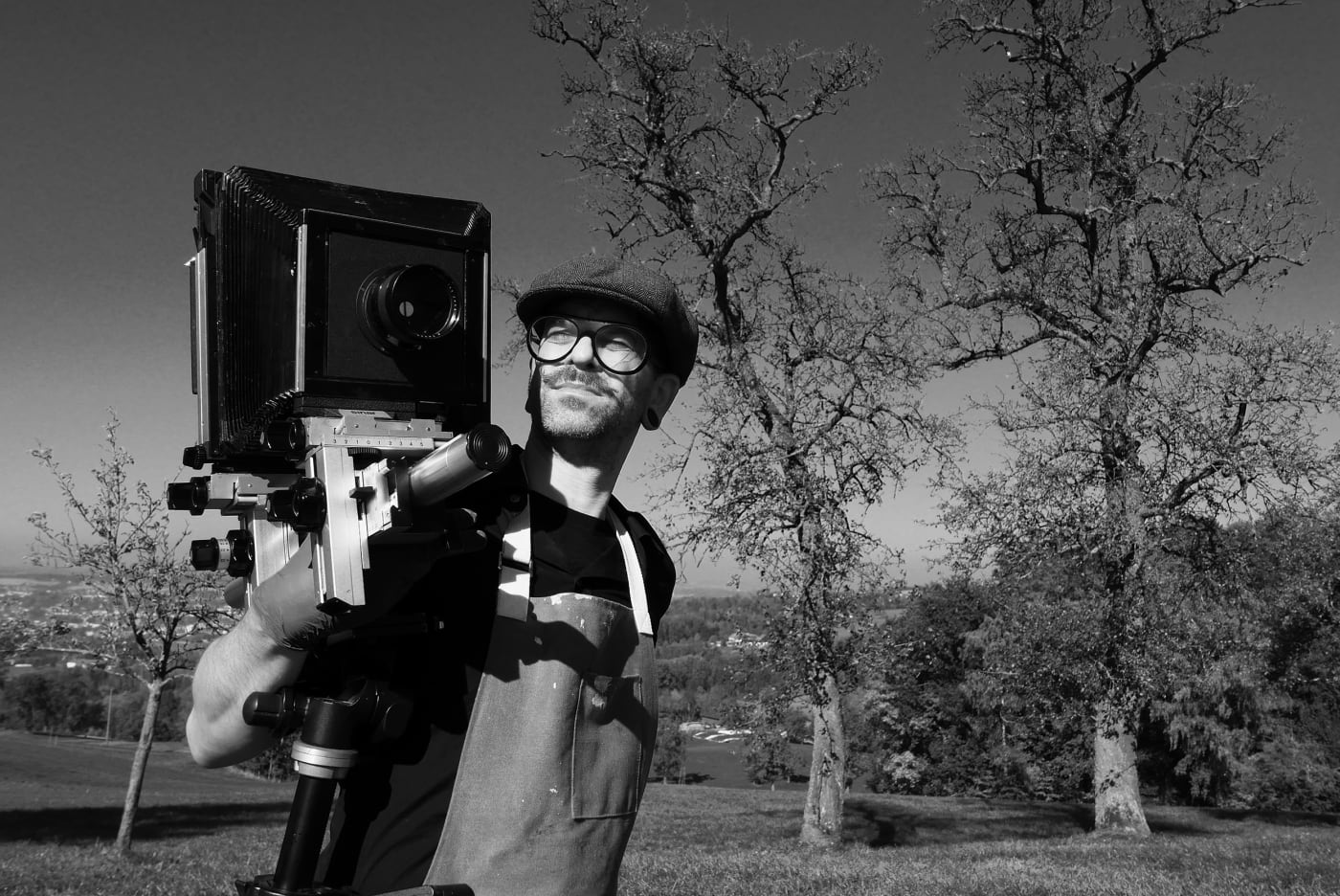 Black and white photo of Rene Huemer holding a large-format camera on a tripod, standing outdoors with leafless trees and a scenic landscape in the background.