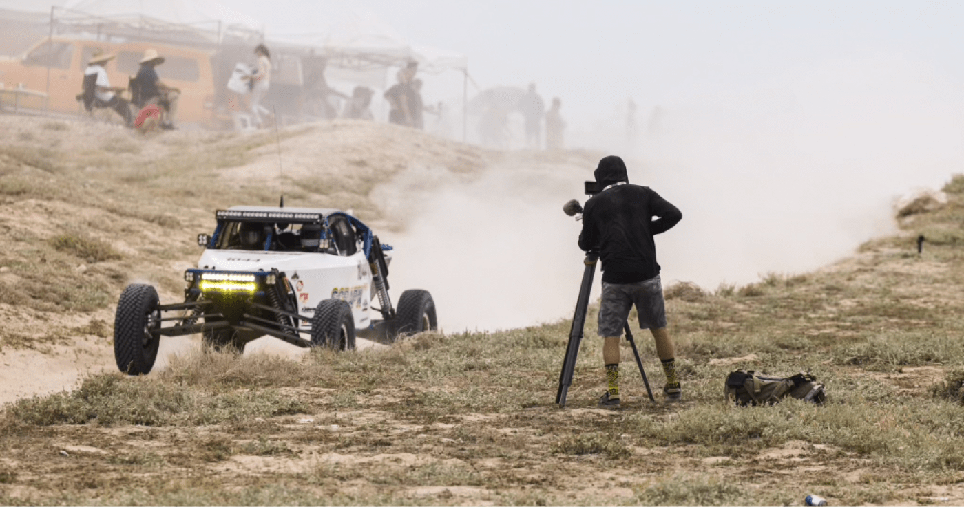 Cahill filming a racing buggy with a tripod on a dusty race track, as spectators watch in the background.