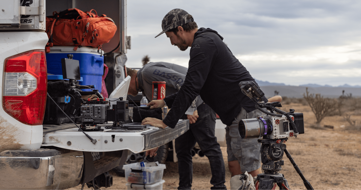 Cahill setting up video equipment on the tailgate of a truck in a desert landscape, with a professional camera on a tripod and gear bags in the background.