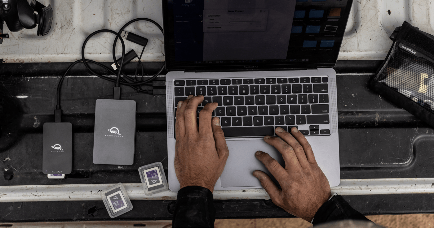 Person working on a MacBook Air connected to OWC Envoy Pro SX and Atlas FXR drives, with memory cards and camera gear on a workspace.