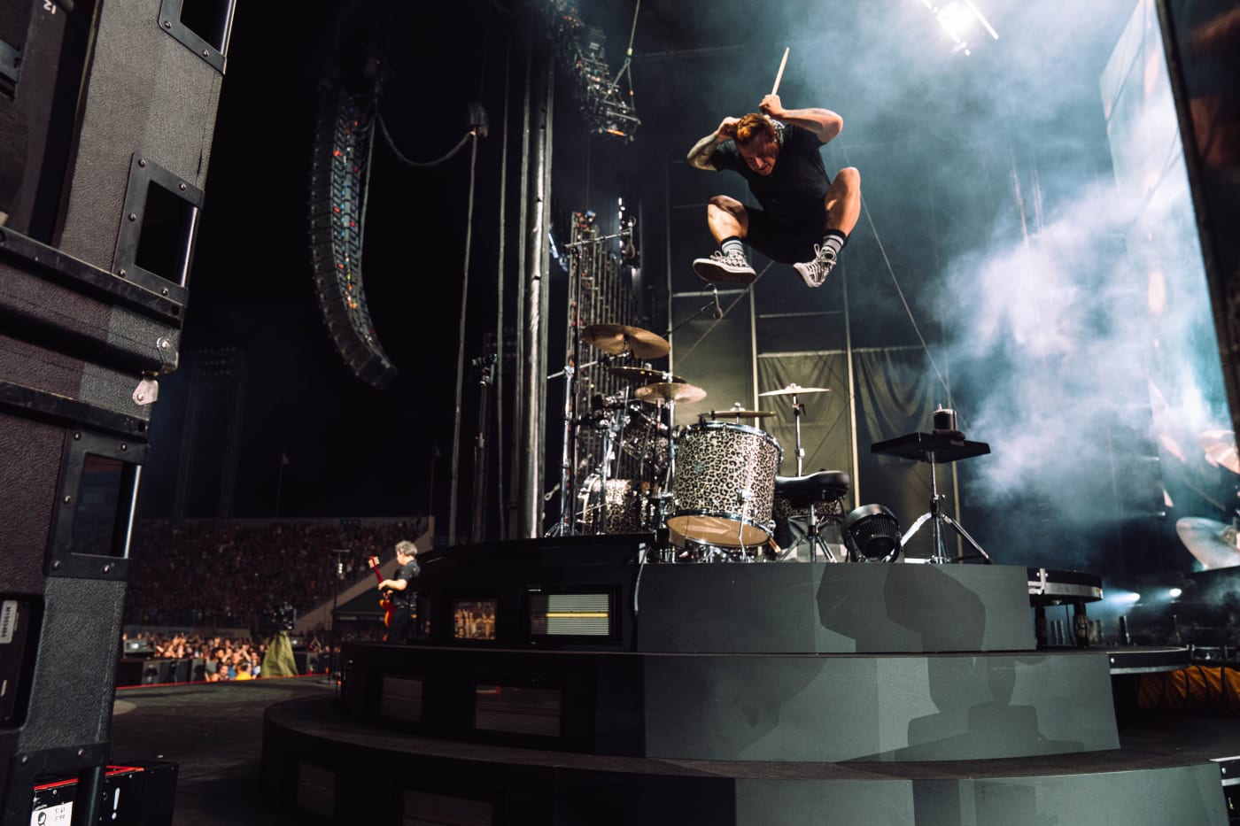 A drummer jumps high in the air while playing drums on stage, captured mid-performance with smoke and stage lights in the background.