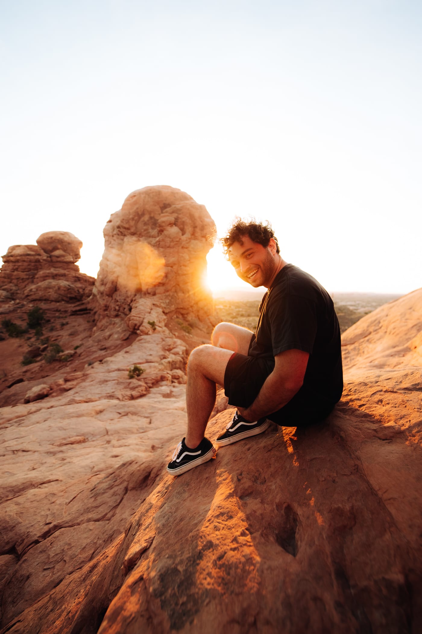 A man smiles while sitting on a rocky ledge at sunset, the warm golden light illuminating the landscape around him.