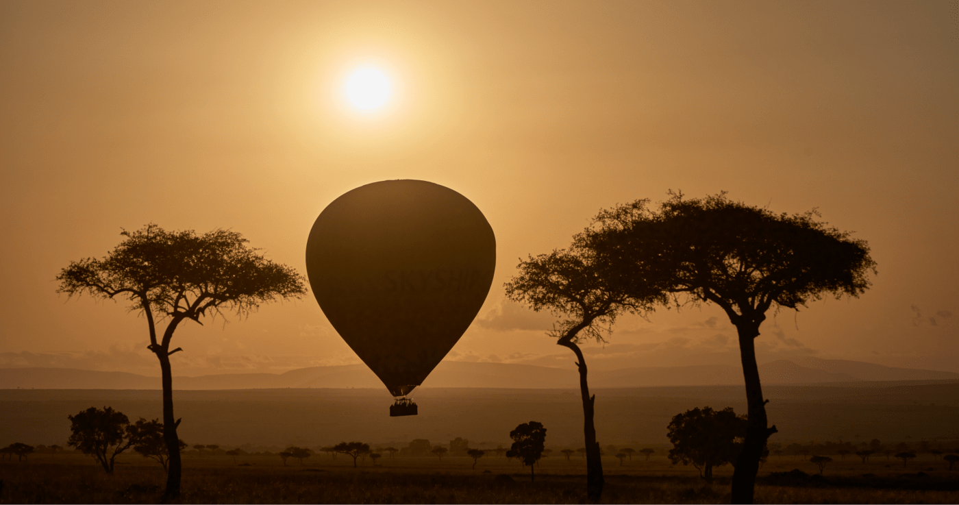 Silhouette of a hot air balloon with the text 'SKYSHIP' floating at sunset between two trees in a savanna landscape, photographed by McNally.