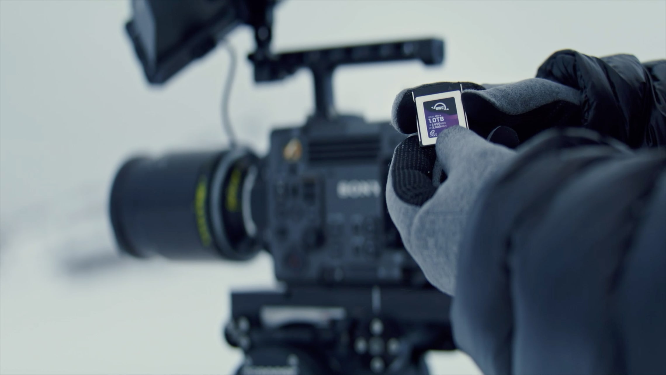 A gloved hand holding an OWC Atlas memory card in front of a professional Sony video camera in a snowy environment.