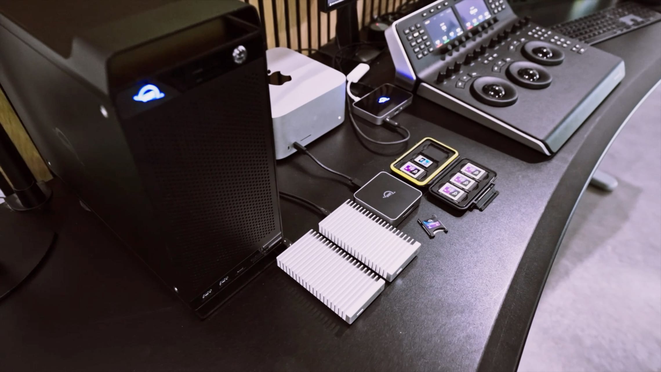 A workstation setup featuring OWC storage solutions, memory cards, a Mac mini, and a professional video editing console.