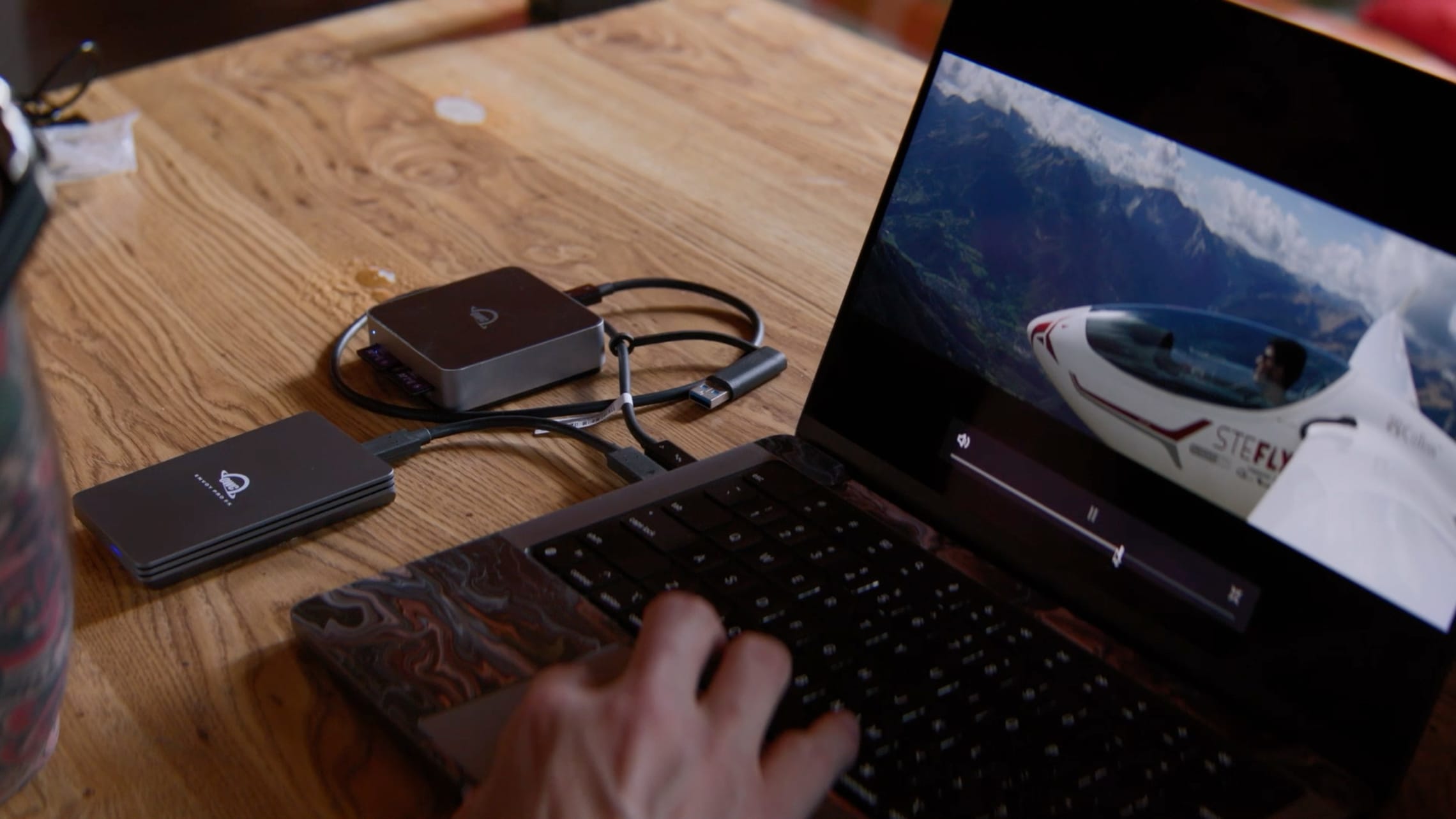 A person working on a laptop connected to OWC storage devices, displaying a high-resolution video of a glider flying over mountains.
        loading=