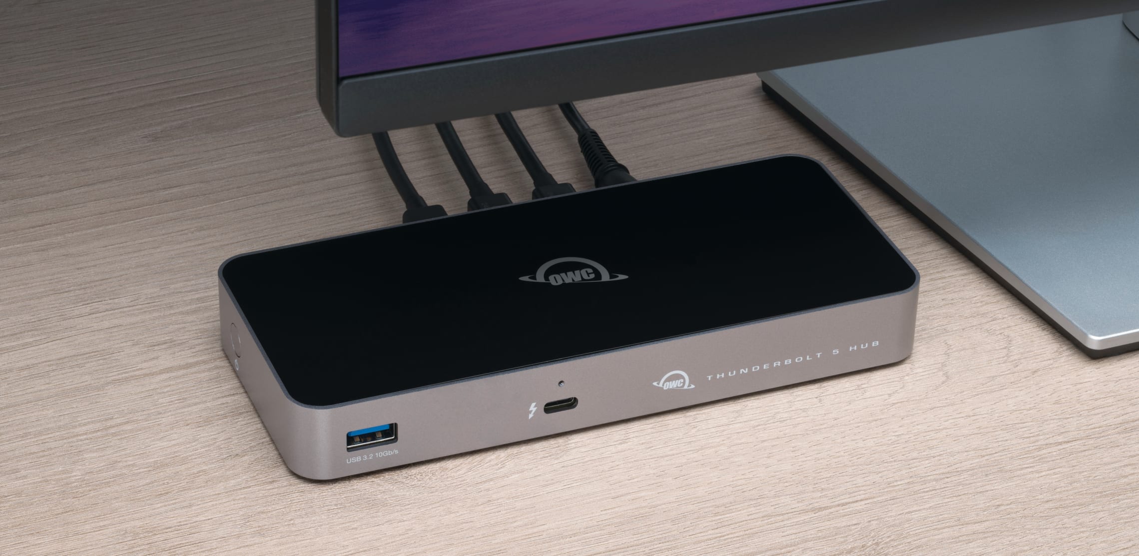 Close-up of the OWC Thunderbolt 5 Hub on a wooden desk, connected to a monitor and showcasing multiple Thunderbolt ports in use, demonstrating high-speed connectivity and device compatibility.