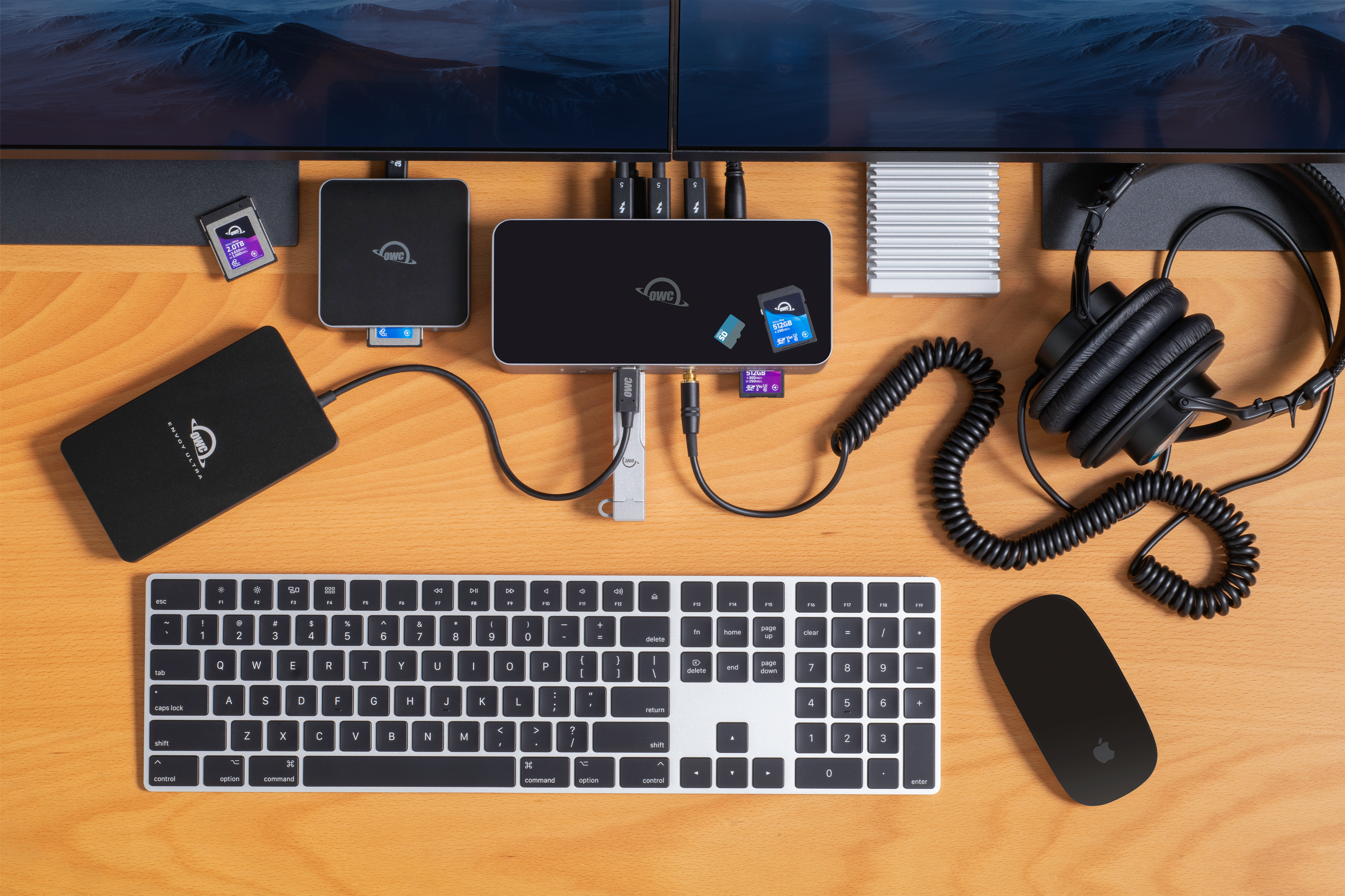 OWC Thunderbolt 5 Dock on desktop with OWC Envoy Ultra, OWC Envoy Pro mini, OWC Express 1M2, OWC Atlas Memory Cards & Card Reader, Apple keyboard and mouse, headphones, and dual displays